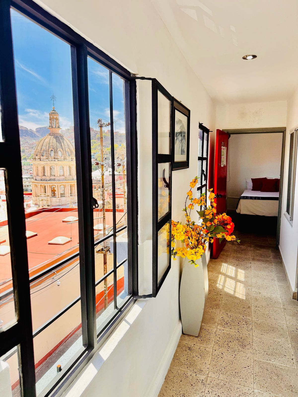 A well-lit hallway is featured, showcasing large windows framing a view of the adjacent building's dome and rooftops. A decorative vase with yellow flowers adds a touch of color. A doorway leads to an inviting bedroom space, visible at the end of the hall.