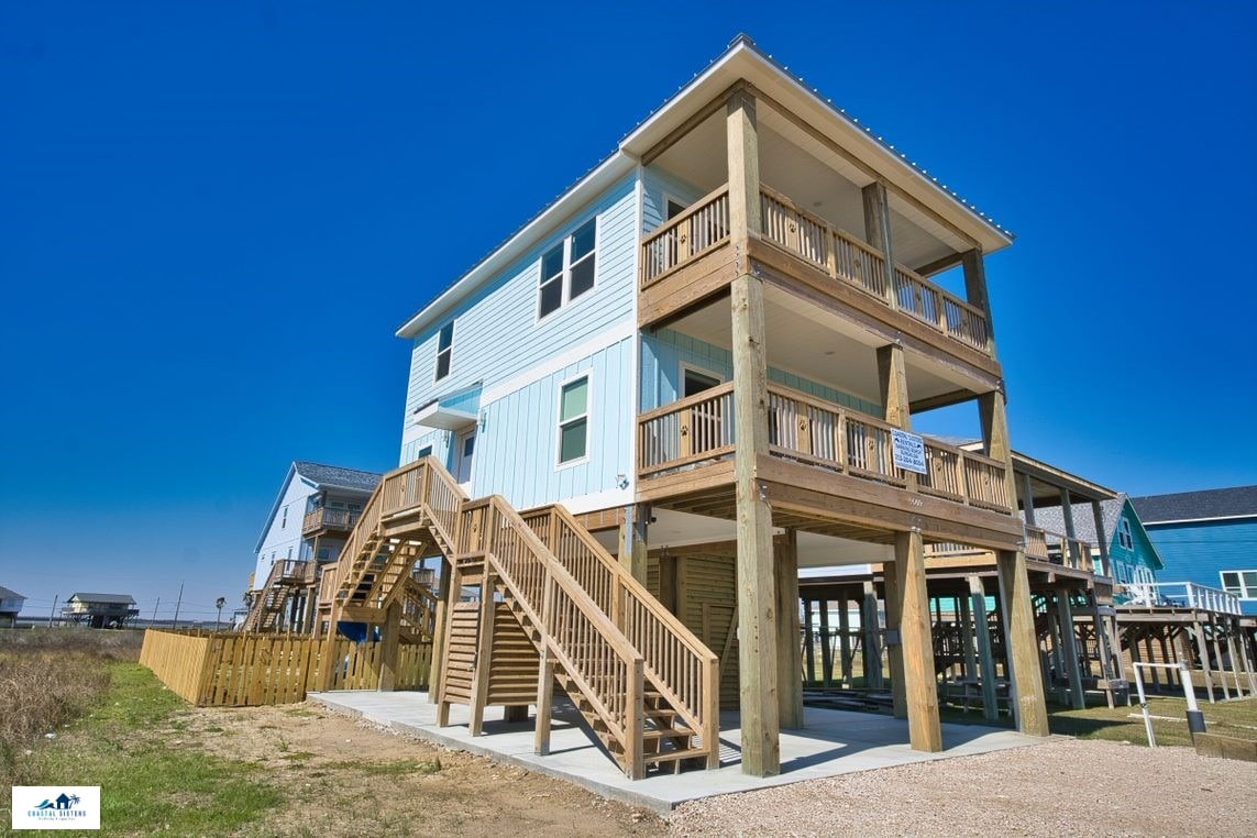 The exterior of a two-story beach house is featured, showcasing a light blue facade with white trim. Multiple decks and stairways made of wood are visible, surrounded by a grassy area. Nearby structures can be seen in the background against a clear blue sky.