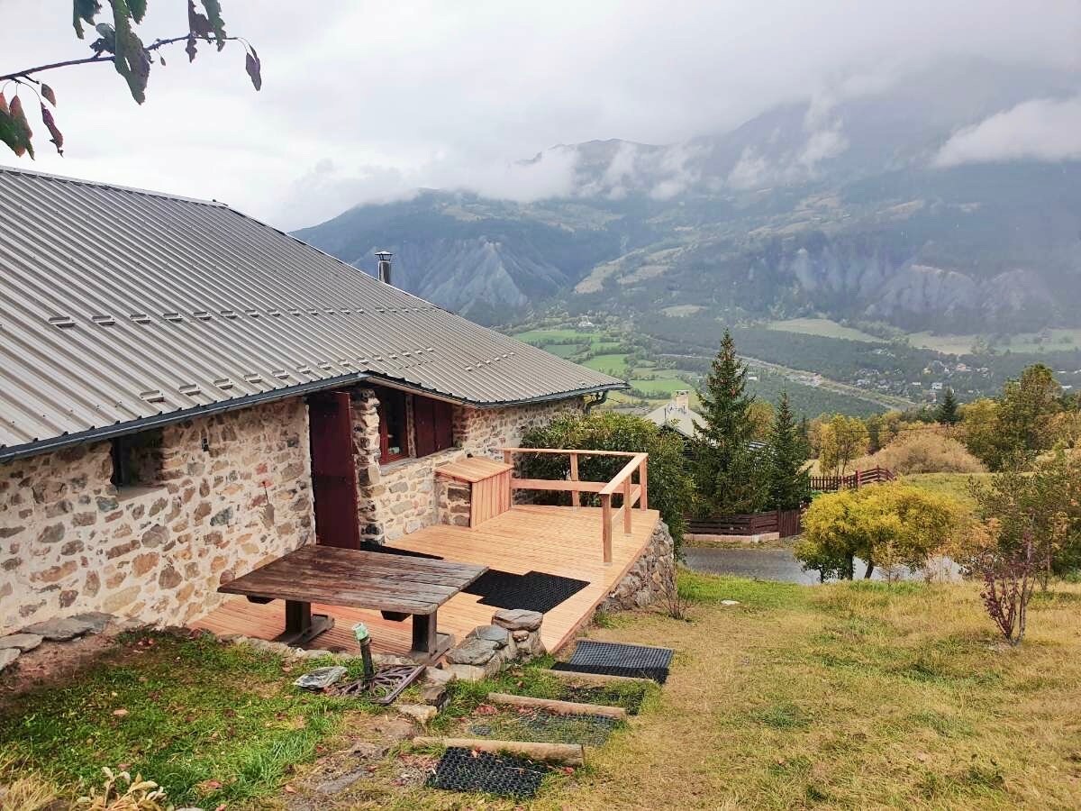 A charming stone house is depicted with a wooden deck overlooking a valley framed by mountains. The outdoor area features a wooden table and benches, with a well-maintained lawn and shrubs surrounding the property. A peaceful and scenic landscape enhances the inviting exterior.