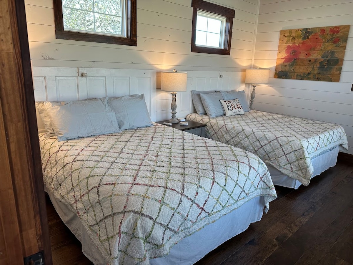 The bright sleeping area features two spacious beds covered with light-colored quilts, arranged beside decorative bedside lamps. Natural light enters through two window panes, illuminating the warm wooden floors and a colorful wall art piece that adds a touch of charm to the room.