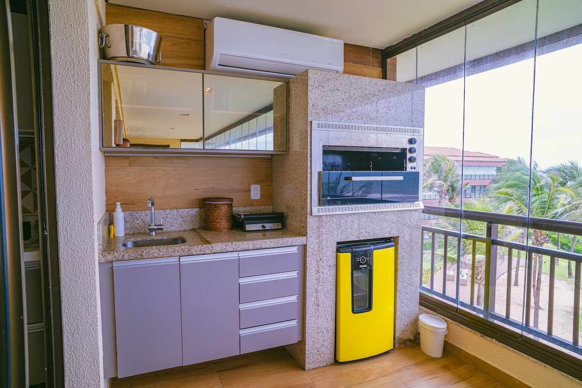 A modern kitchenette is showcased, featuring sleek cabinets in muted grey and a bright yellow refrigerator. A microwave is positioned above the counter, with a sink and a decorative basket on the counter. Large windows allow natural light and views of the surrounding greenery.