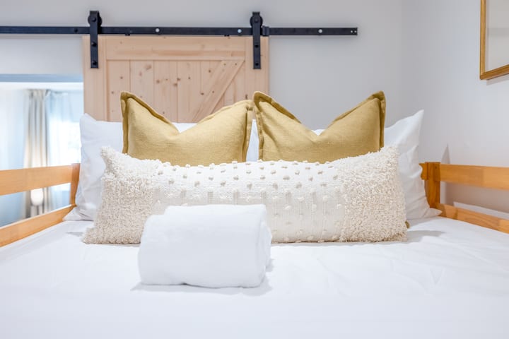 צילום תקריב של מיטת הקומתיים העליונה המעוצבת עם כריות מפוארות בגוונים ניטרליים, מרקמים רכים ושתי מגבות מגולגלות יוצר סידור מזמין. מעקות עץ לבטיחות ודלת האסם הזזה ברקע מוסיפים מגע חמים וכפרי.