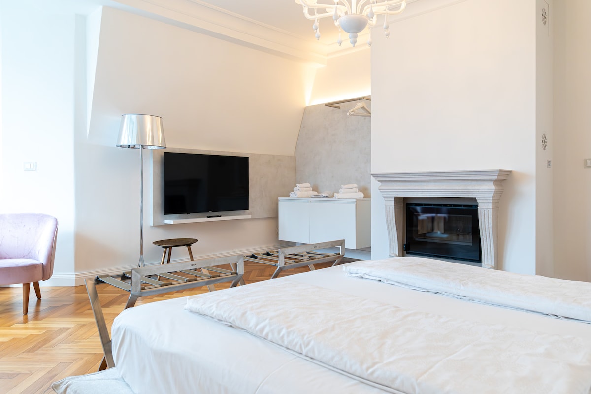A well-appointed room features a spacious bed with soft linens. A flat-screen TV is mounted on the wall, complemented by a modern fireplace. A stylish armchair and a small table enhance the seating area, while neatly arranged towels add a touch of convenience.