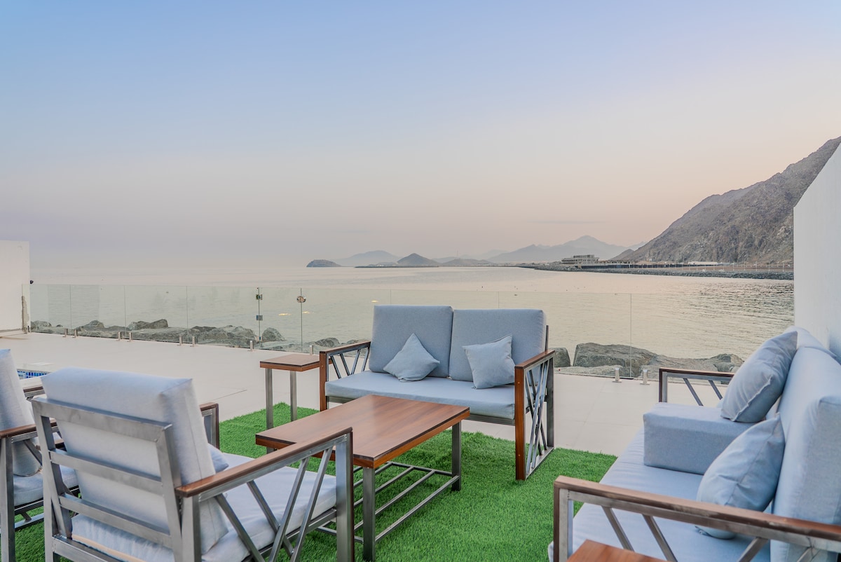 An outdoor lounge area features comfortable seating with soft cushions and wooden frames, arranged around a small wooden table. The seating overlooks the tranquil sea, with mountains visible in the background. The area is partially covered by modern glass railings, enhancing the view.