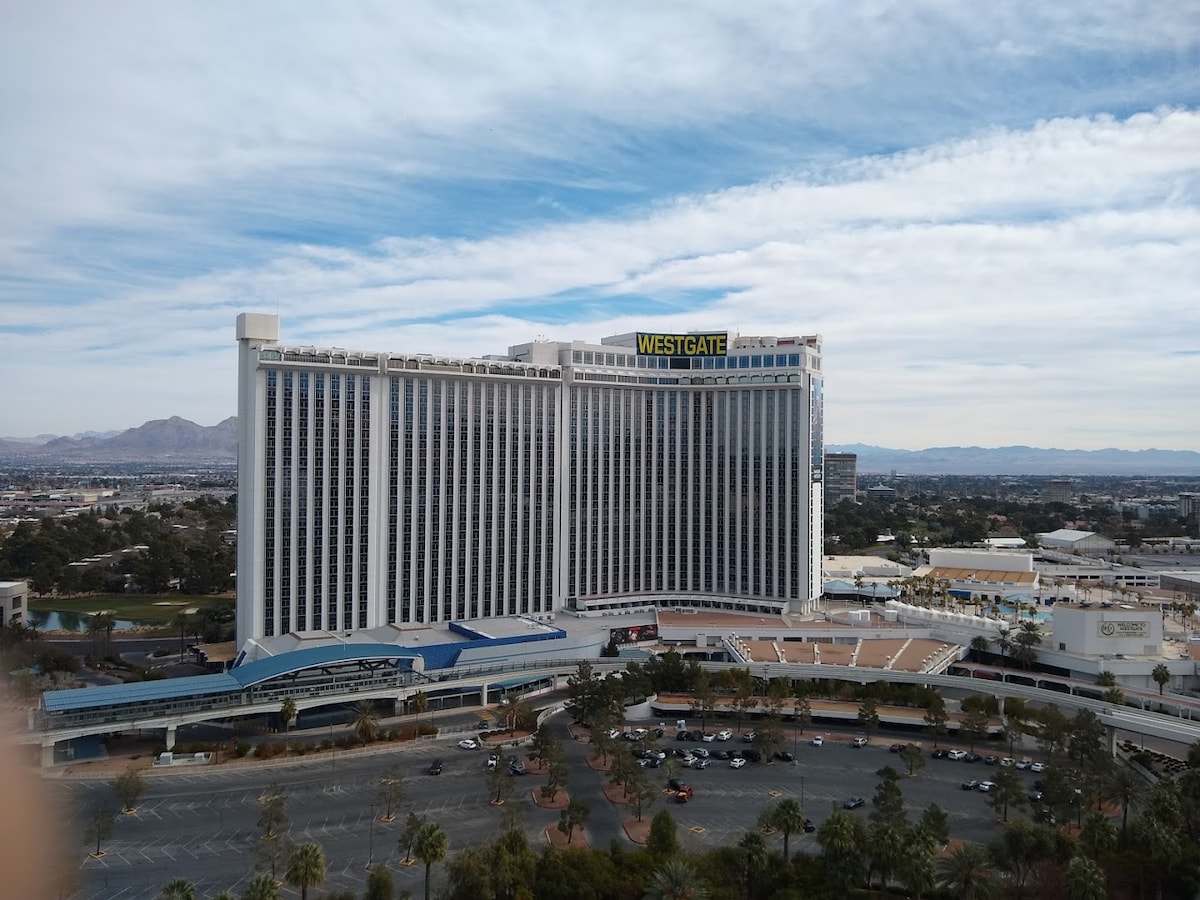 The Westgate Las Vegas Resort & Casino is prominently featured with its tall, modern façade. Surrounding areas include landscaped pathways and parking spaces. The backdrop showcases distant mountains under a partly cloudy sky, providing a glimpse of the expansive Las Vegas landscape.