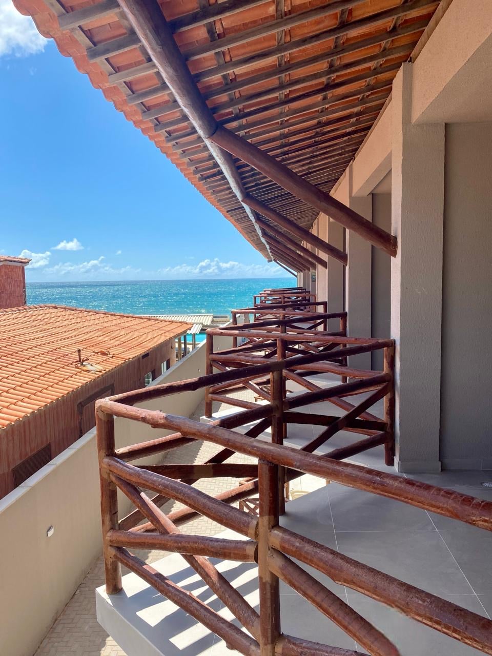 A spacious balcony area is framed by rustic wooden railings, overlooking a vibrant seascape. The shoreline and blue waters are visible in the distance, complemented by a clear sky. The architecture showcases a blend of natural materials with a coastal ambiance.