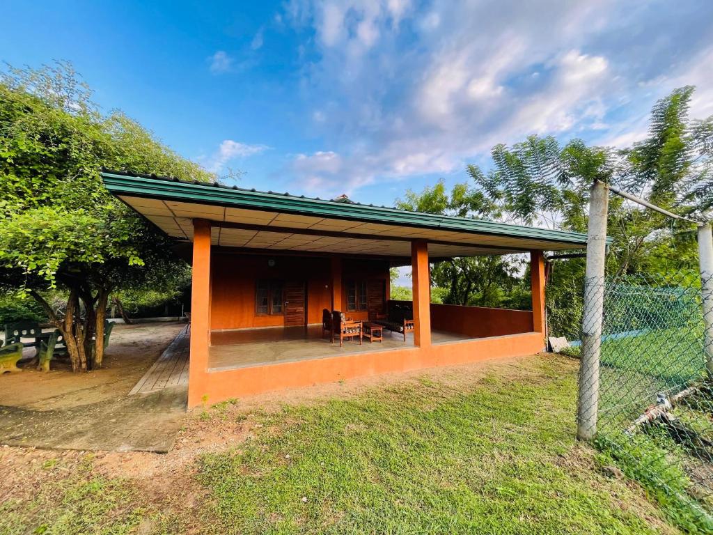 A covered outdoor patio features wooden seating arrangements, providing a relaxed space to enjoy nature. Lush greenery surrounds the area, contributing to a tranquil setting. An open grassy area is visible beyond the patio, with trees adding depth to the landscape.