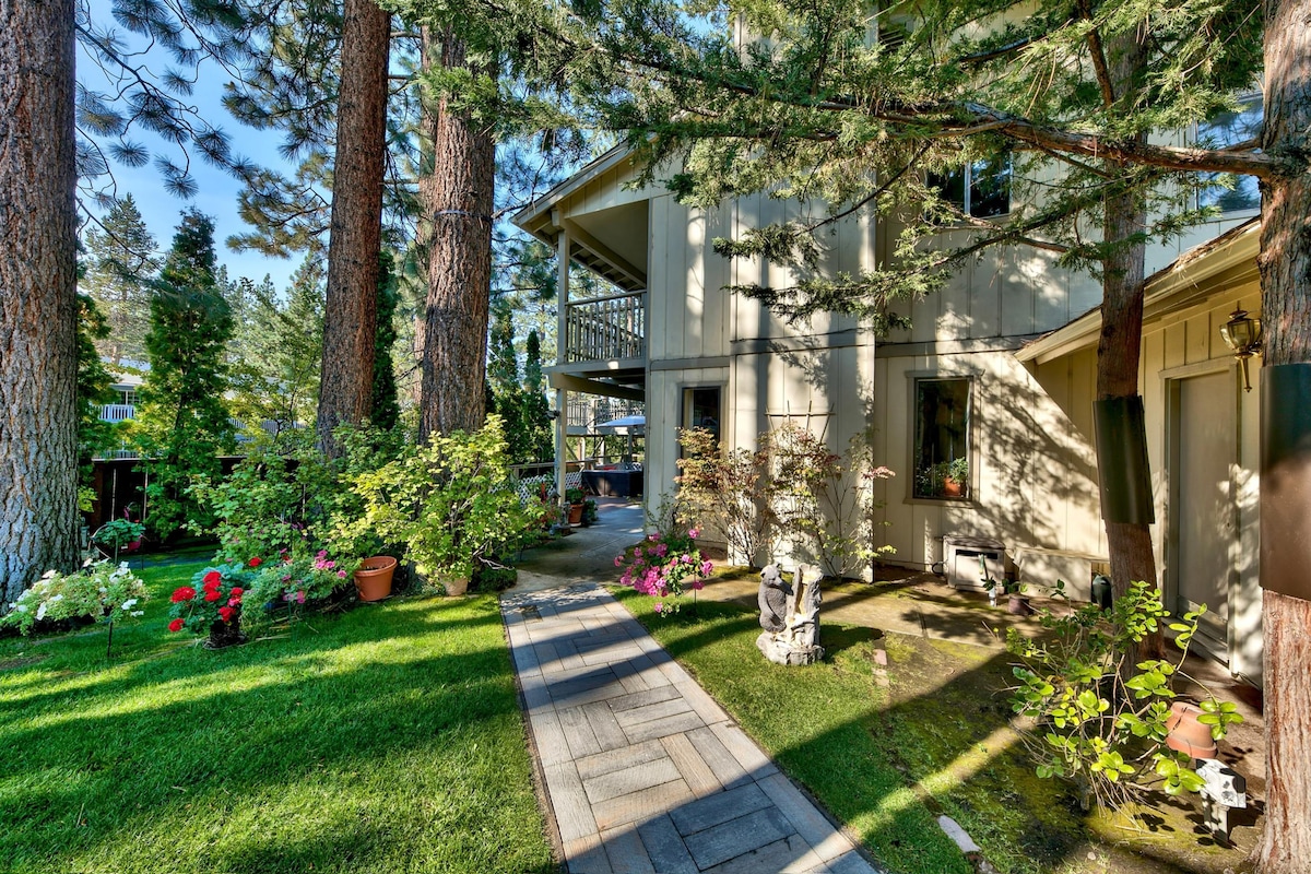 A paved walkway leads alongside a well-maintained yard, featuring vibrant flower beds and lush greenery. Tall trees provide shade nearby, contributing to a naturally serene setting. The exterior of the house showcases a blend of light-colored siding and multiple levels, offering ample outdoor space.