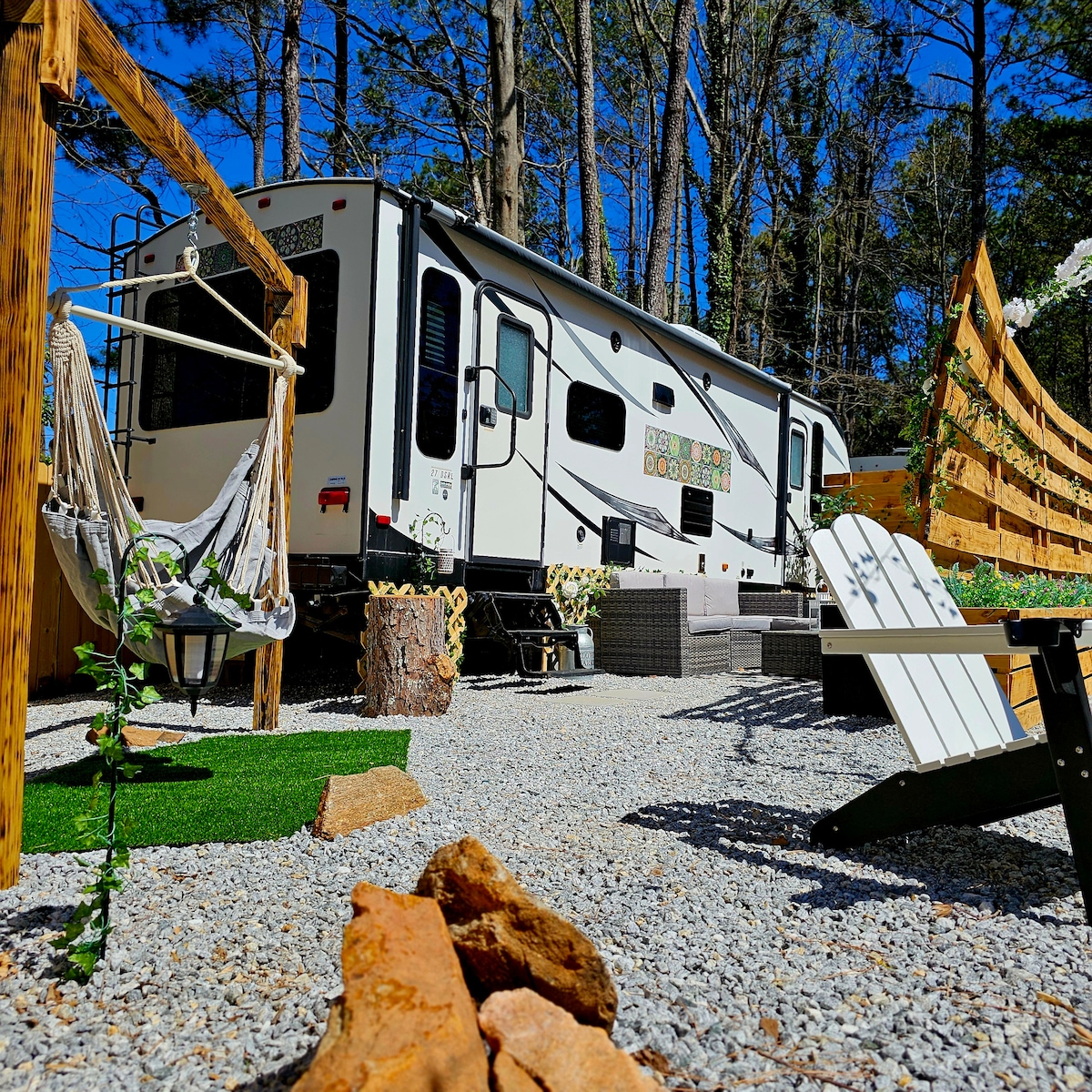 A charming glamper is nestled among trees, surrounded by pebbles and greenery. A hammock chair hangs nearby, while seating areas are arranged using wooden furniture. Natural sunlight brightens the outdoor space, enhancing the inviting atmosphere of the glampsite.