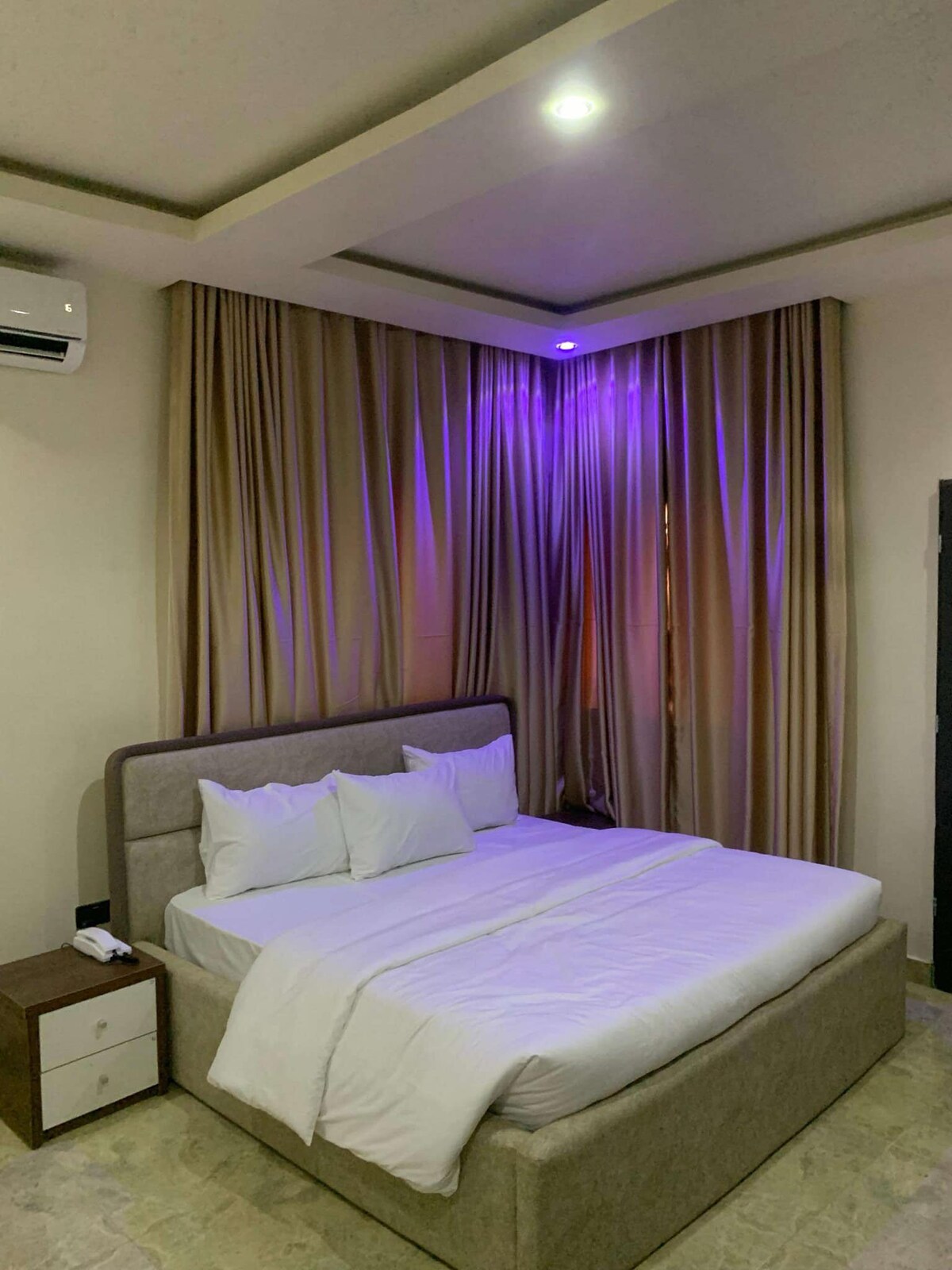 A spacious bed with crisp white linens is positioned against a softly lit wall. Floor-to-ceiling curtains frame the window, emitting a gentle purple hue from integrated lighting. A bedside table is visible, enhancing the room's functionality.