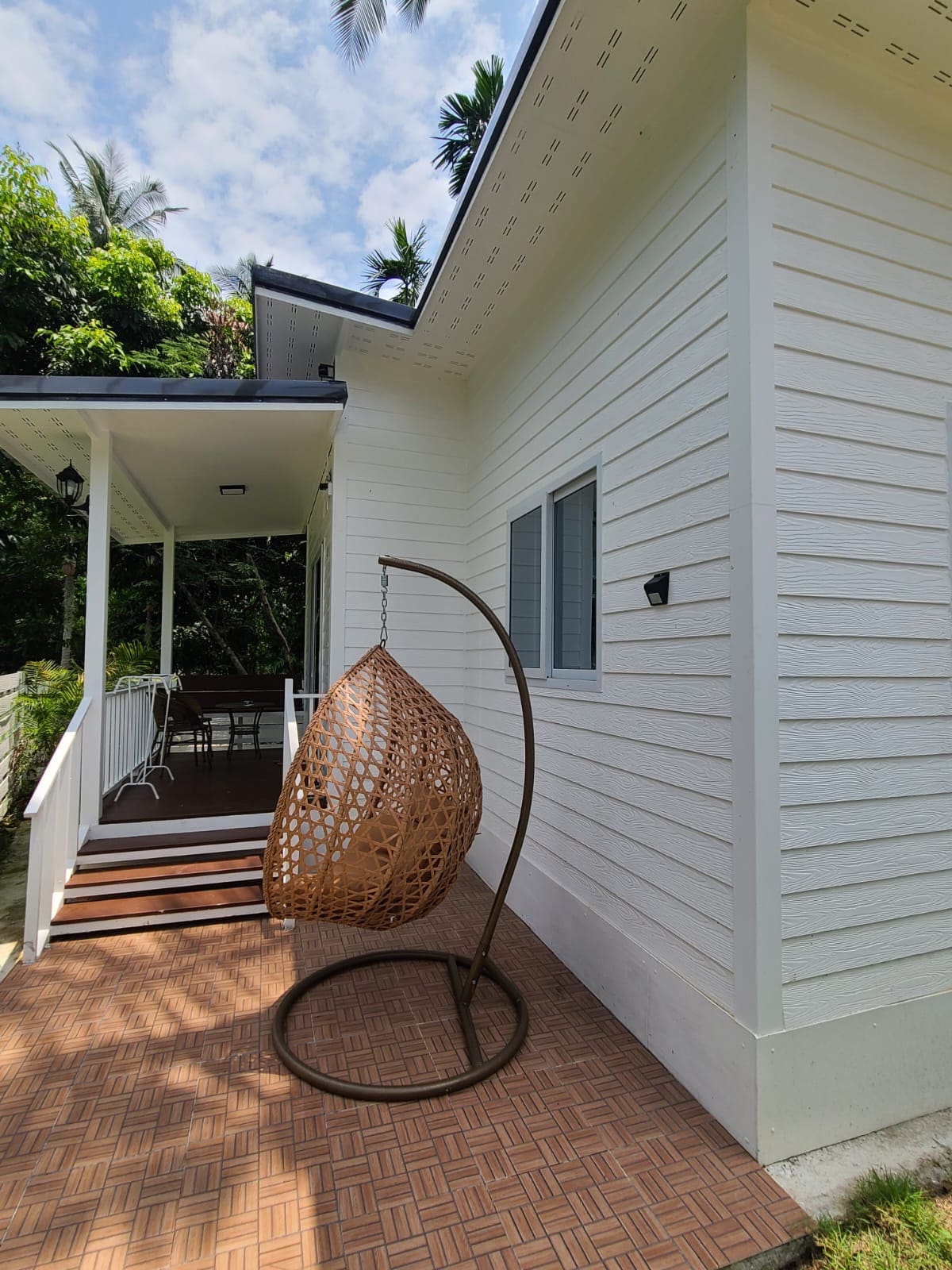 An outdoor seating area features a stylish wicker hanging chair on a patterned tile surface. The white bungalow exterior is complemented by a small porch, and a large window offers a glimpse of the interior. Lush greenery surrounds the space, enhancing the natural setting.