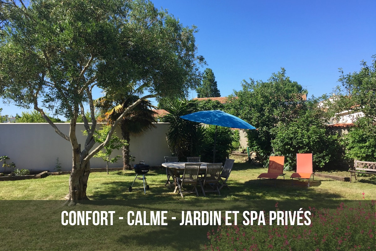 A spacious garden is shown, featuring a shaded dining area with a table and chairs, surrounded by lush greenery and trees. Two sun loungers are positioned on the grass, and a blue umbrella provides additional shade. The private spa area is visible, inviting relaxation.