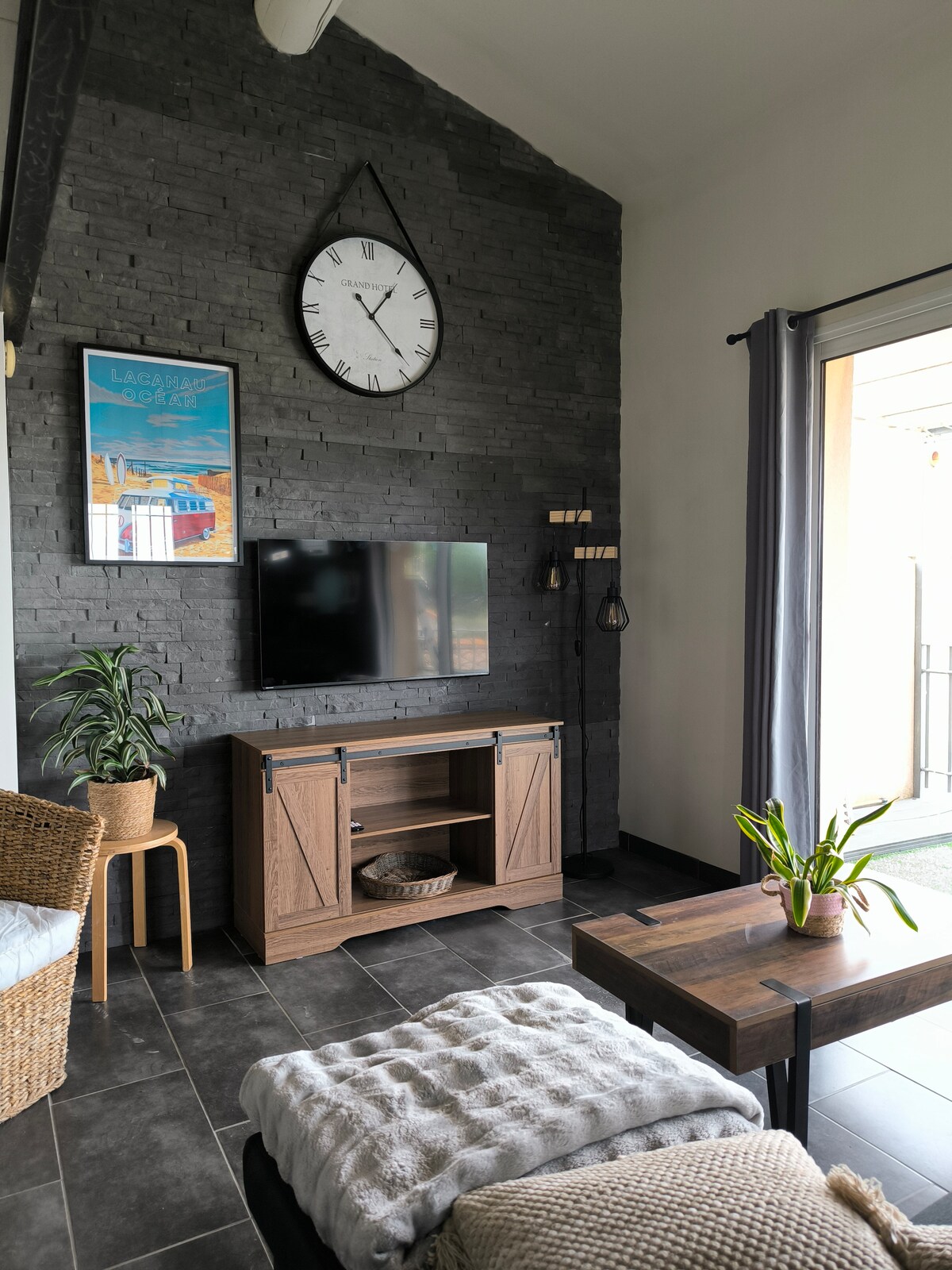 A living area features a blend of modern and rustic elements, including a dark stone wall adorned with a large clock and artwork. A flat-screen television is mounted above a wooden cabinet. A potted plant and a decorative throw add warmth to the space.
