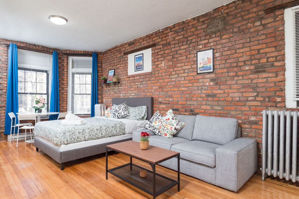 A studio apartment features exposed brick walls and natural wood flooring, complemented by large windows with blue curtains. A spacious bed with decorative pillows is positioned next to a minimalist gray sofa. A coffee table sits in front of the seating area, enhancing the open layout.