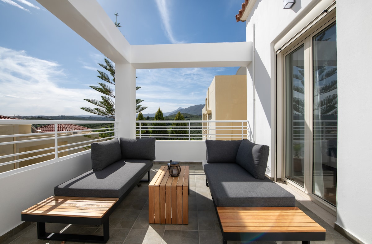 An inviting balcony features two spacious lounge sofas positioned around a wooden coffee table. The area is framed by railings and is complemented by a clear sky and distant mountains, providing a serene outdoor setting for relaxation.