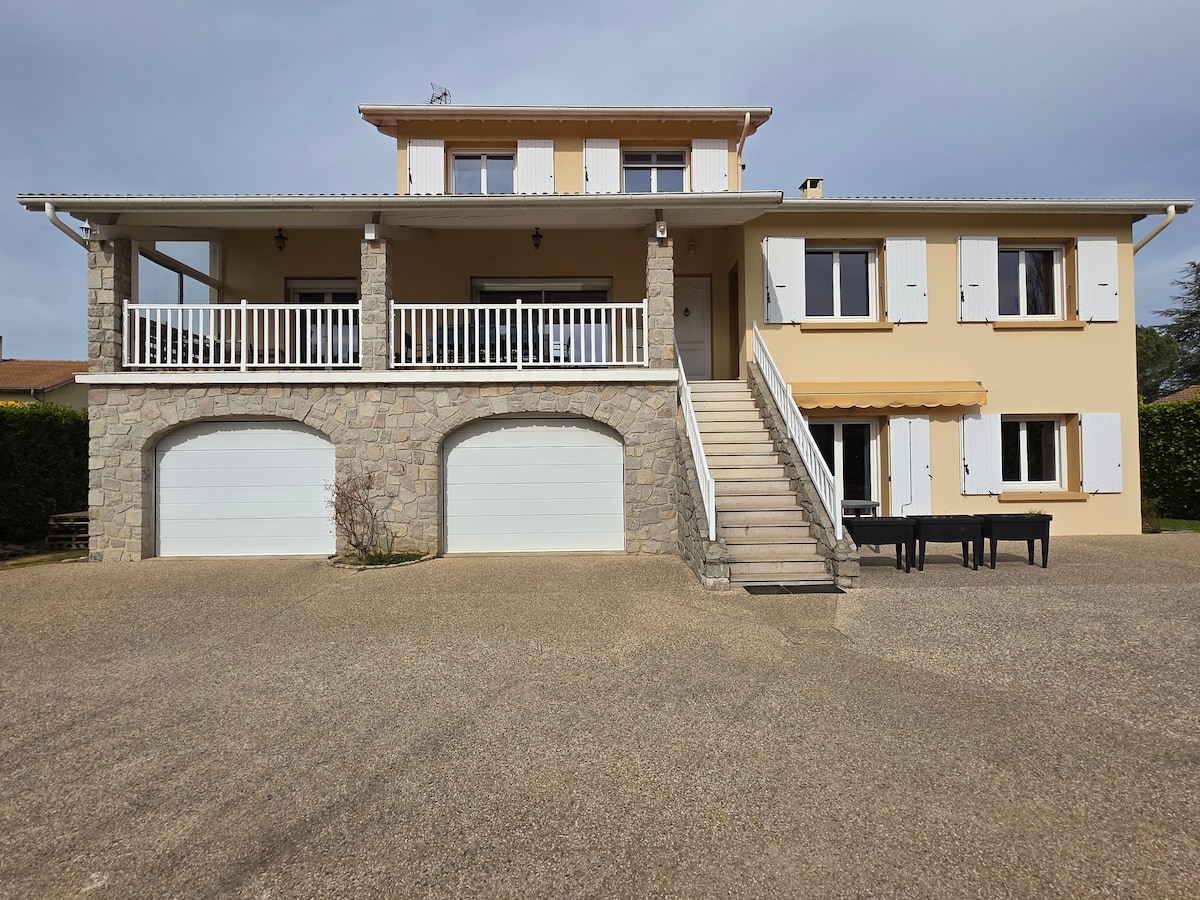 A two-story home features a light-colored exterior with white shutters. A covered porch is supported by stone columns, leading to a set of stairs. Two garage doors are visible below, and a large outdoor space is evident in front of the building.