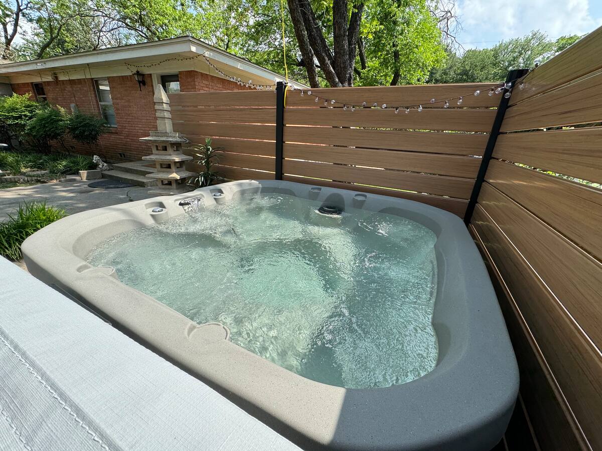 A spacious hot tub is surrounded by a wooden privacy fence, featuring jets that create gentle bubbles within the clear water. Natural sunlight filters through nearby trees, highlighting the inviting atmosphere of this relaxing outdoor space.