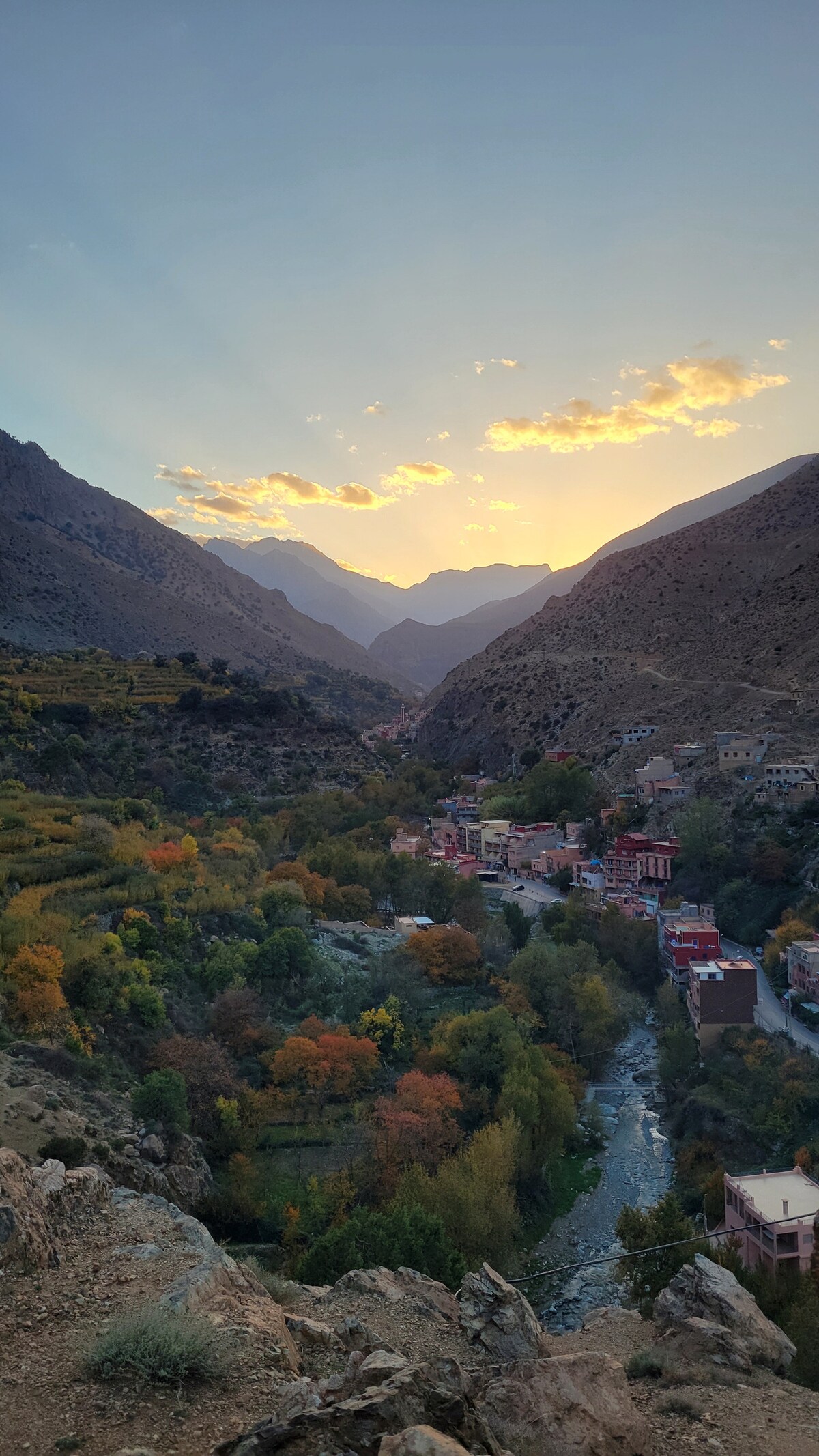 A panoramic view captures a valley flanked by steep mountains, with a winding river below. The scene is adorned with lush greenery and trees displaying autumn hues, while a small village is nestled in the valley, illuminated by warm sunset tones in the sky.
