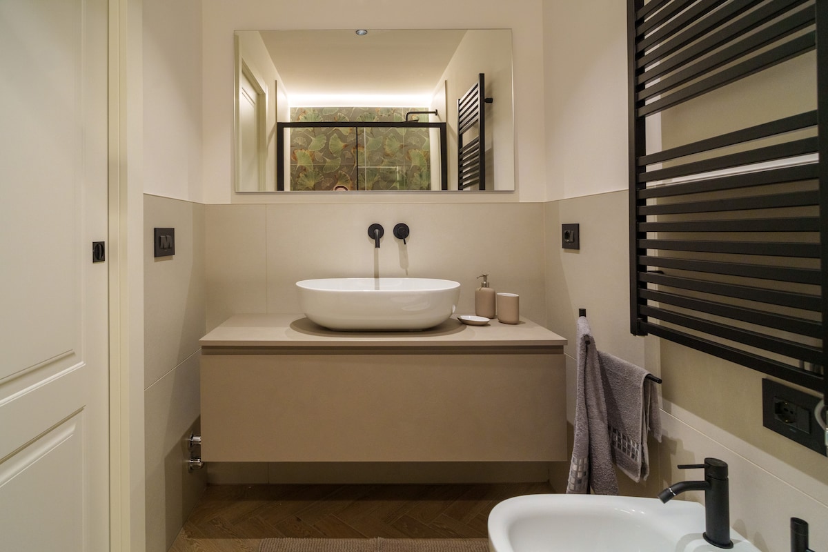 A modern bathroom features a stylish countertop basin under a wide mirror. Wall-mounted fixtures are positioned above a sleek storage unit, with a towel hanging nearby. A white bidet and a dark-framed towel radiator complete the space, creating a practical yet elegant environment.