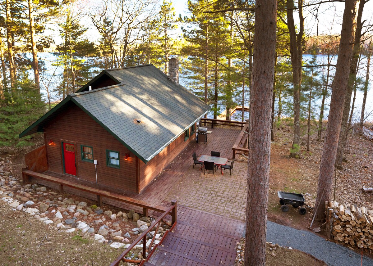 A wooden cabin is situated among tall trees, with a large deck surrounding it. A stone pathway leads to the cabin entrances. Tables and chairs are positioned on the deck, with a view of the tranquil waters in the background.