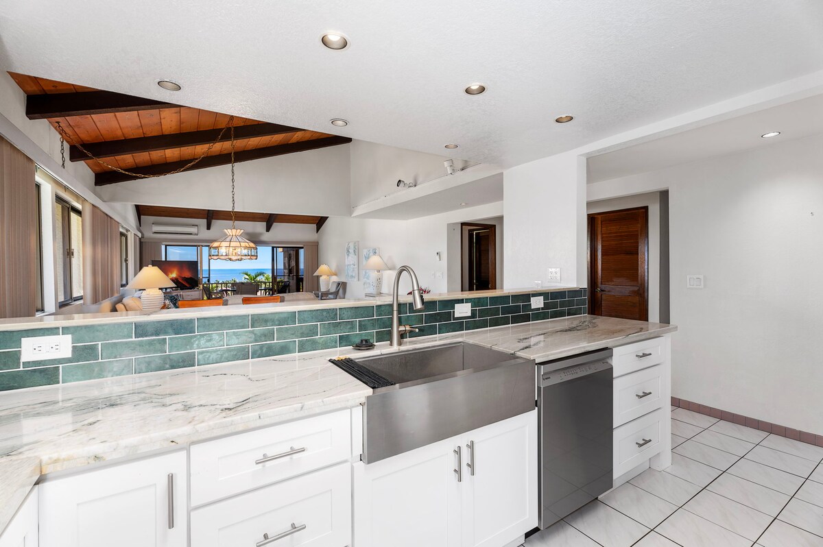 A well-appointed kitchen features modern stainless steel appliances, including a dishwasher and a large sink. Elegant white cabinetry and marbled countertops contrast with green tiled accents. The open design allows for views into the spacious living area, enhanced by natural light from multiple windows.