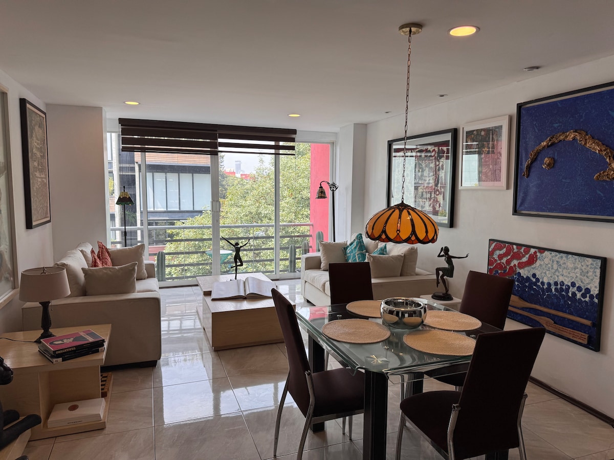 A bright living area features large windows with views of surrounding trees. Two light-colored sofas are arranged with decorative pillows, and a glass dining table is surrounded by dark chairs. Artistic wall decor adds character to the space, complemented by warm lighting from a Tiffany-style lamp.