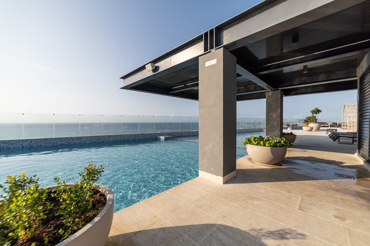 A swimming pool is situated on a rooftop terrace, featuring clear blue water that reflects the sky. The pool area is surrounded by modern planters with greenery. A panoramic view of the ocean is visible beyond the transparent railing, enhancing the open atmosphere.