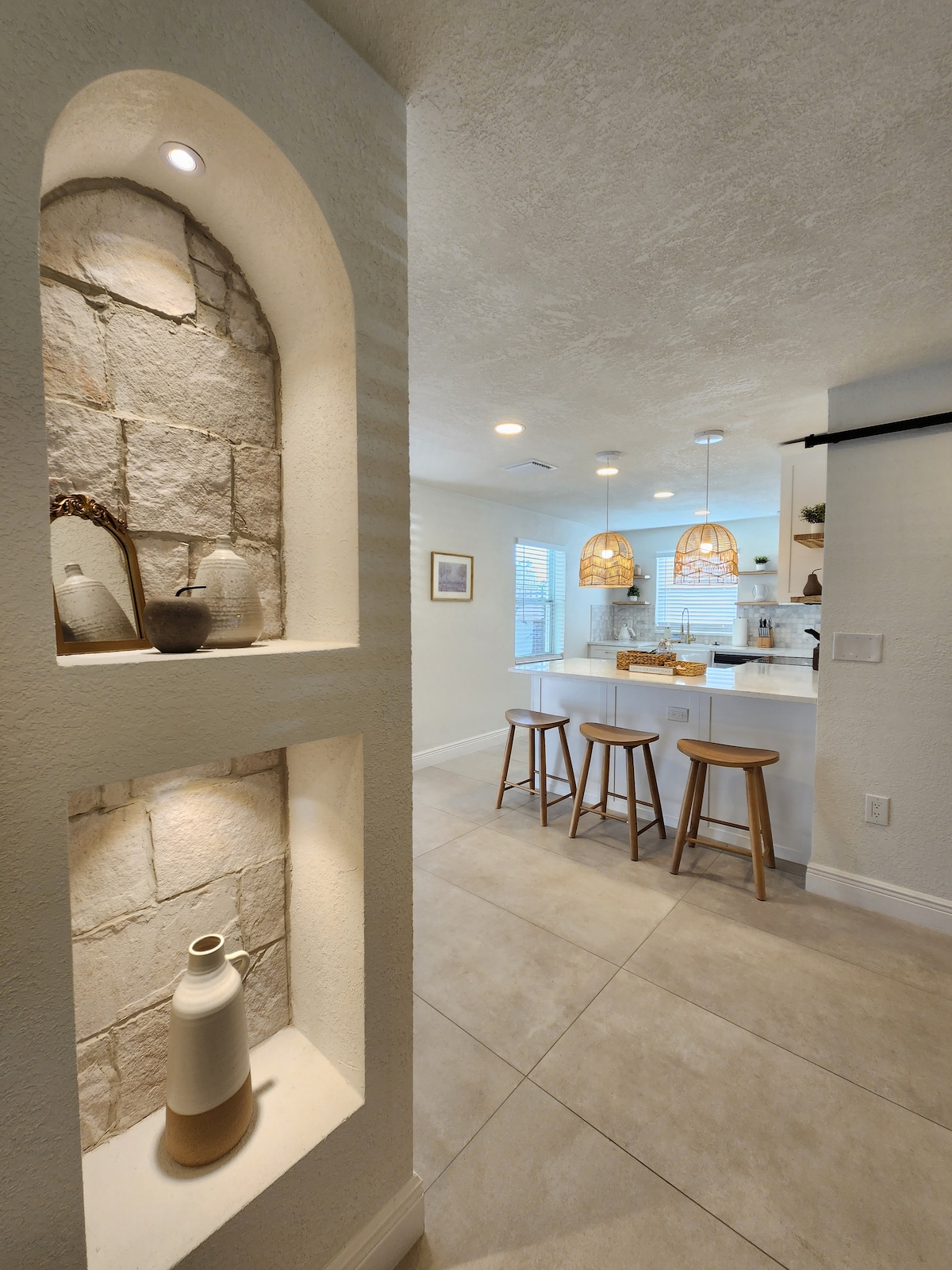 An open kitchen area features modern bar stools positioned around a white island topped with stylish pendant lighting. The adjacent wall showcases a recessed niche displaying ceramic vases, illuminated softly. Natural light filters in through the nearby windows, casting a bright and welcoming glow.