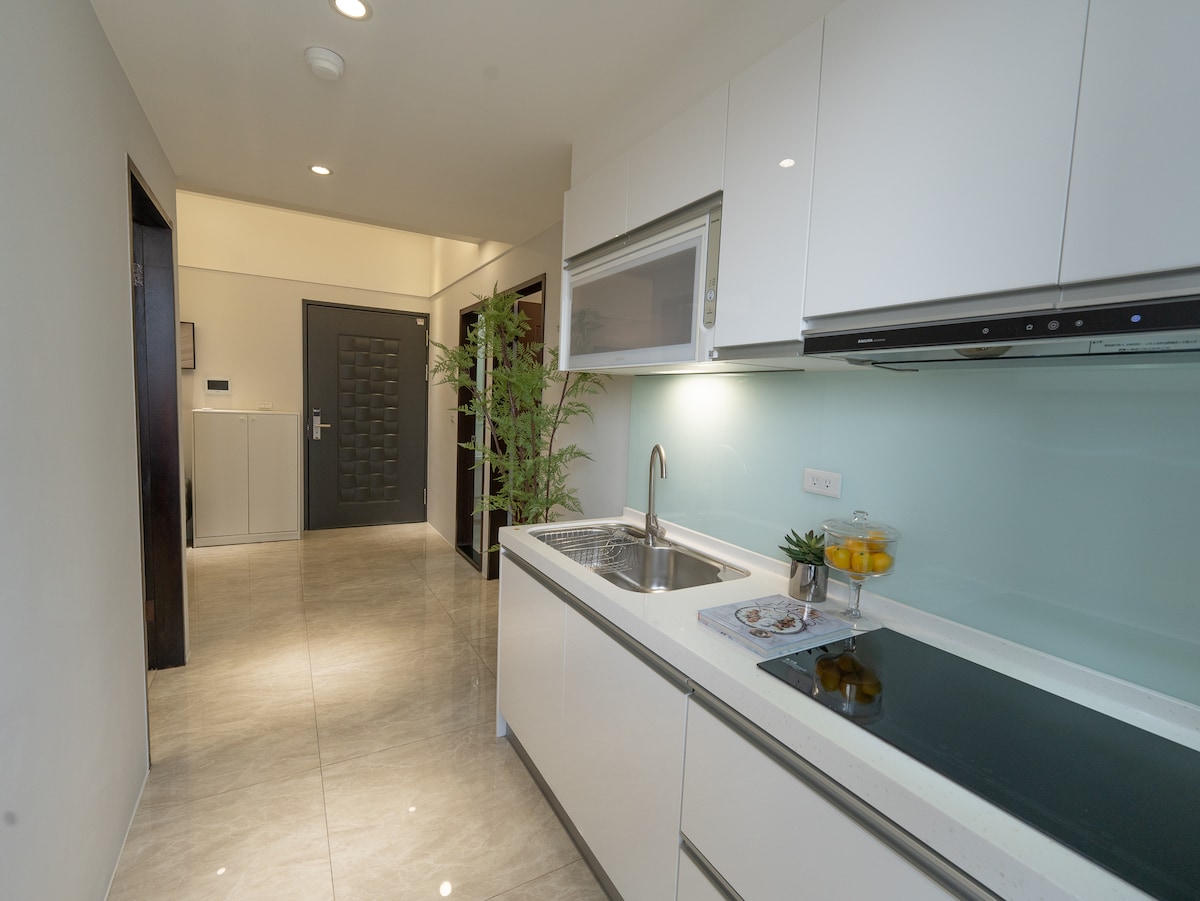 A modern kitchen is presented, featuring sleek white cabinetry and a stainless steel sink. An induction cooktop is visible alongside a microwave. Natural light enhances the bright atmosphere, with a small plant adding a touch of greenery.