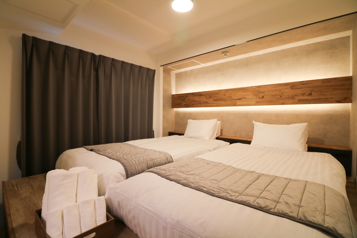 A well-lit sleeping area features two spacious single beds, dressed in white linens and accented with gray blankets. Soft lighting along the wooden wall creates a calming ambiance. A neatly arranged set of towels is placed on a wooden surface beside the beds.