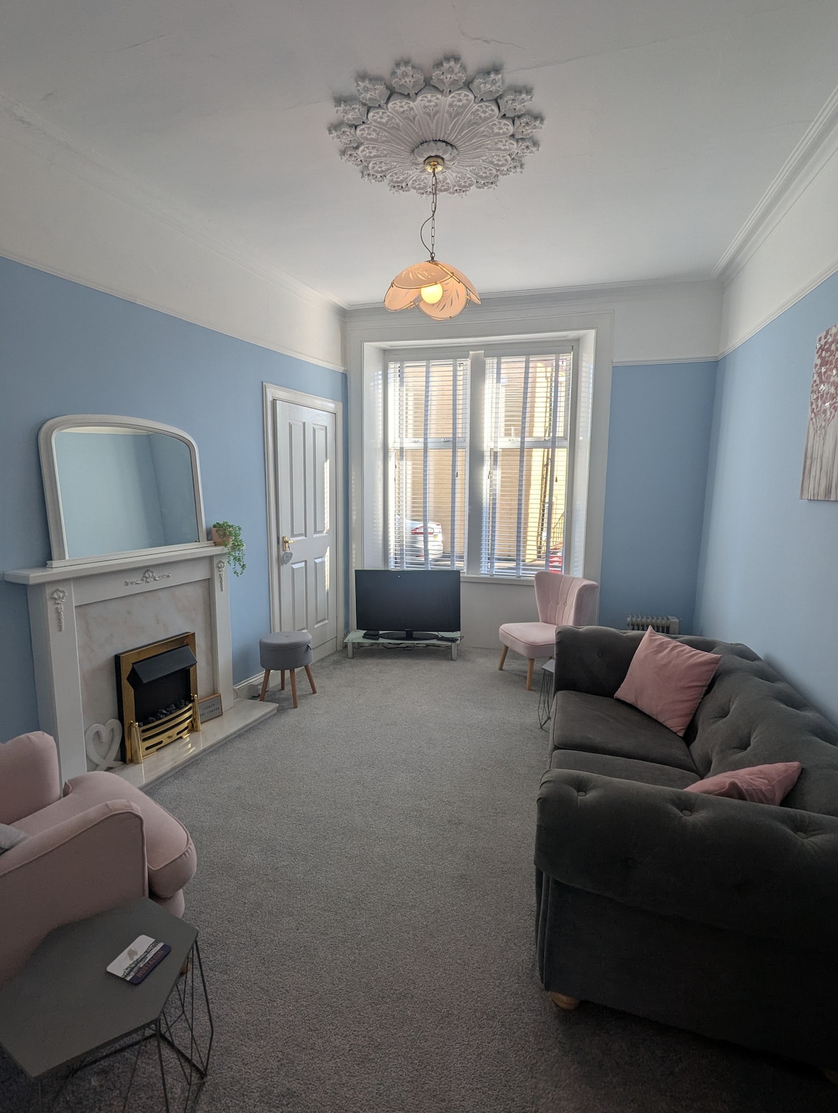 A light blue living room features a cozy grey sofa and two pink armchairs, complemented by a small white coffee table. A modern gas fireplace adds warmth, while a wide-screen TV is positioned across from the seating. Natural light filters through the elegant window treatment.