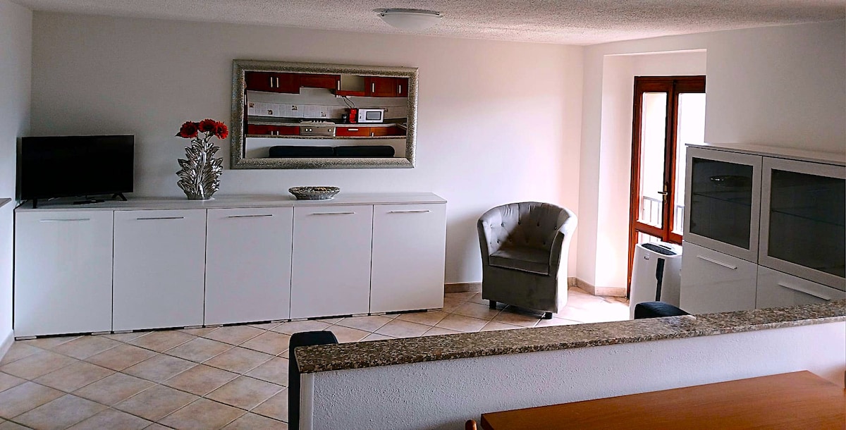 A spacious living area features tiled flooring and a large window allowing natural light. A neutral color palette is complemented by a decorative mirror over a white cabinet. A comfortable chair and television add to the inviting ambiance of the space.