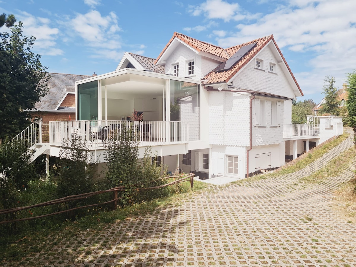 A multi-level house is displayed with a bright white exterior, featuring large windows and a modern glass-enclosed terrace. The structure is surrounded by greenery, with a pathway leading to the entrance. A clear sky and scattered clouds provide a serene backdrop.