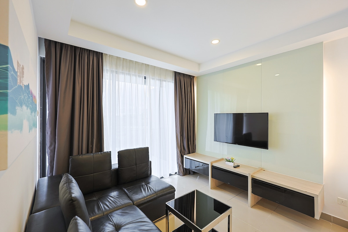 A modern living space features a black leather sectional sofa opposite a flat-screen TV mounted on a light-colored wall. Sunlight filters through sheer curtains, illuminating the area, while a square black coffee table sits in the center, enhancing the open layout.
