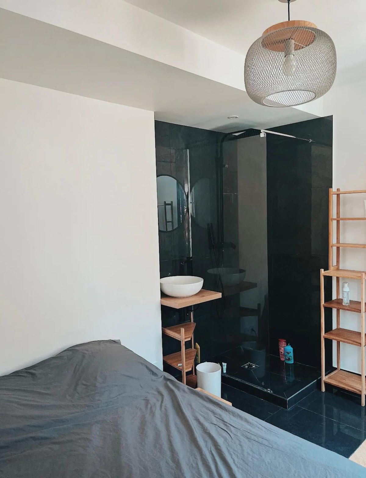 A modern sleeping area features a spacious bed with a neutral grey cover. A sleek shower area is visible, enclosed in dark tiles. A stylish hanging lamp provides soft illumination, while a wooden shelf stands nearby, holding toiletries and decorative items.