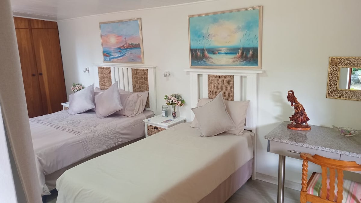 A spacious bedroom is highlighted by a double bed and a single bed, both adorned with soft, neutral bedding. Decorative wall art frames two landscapes above each bed, while a small desk provides additional workspace. Natural light fills the room, complemented by light-colored furnishings.