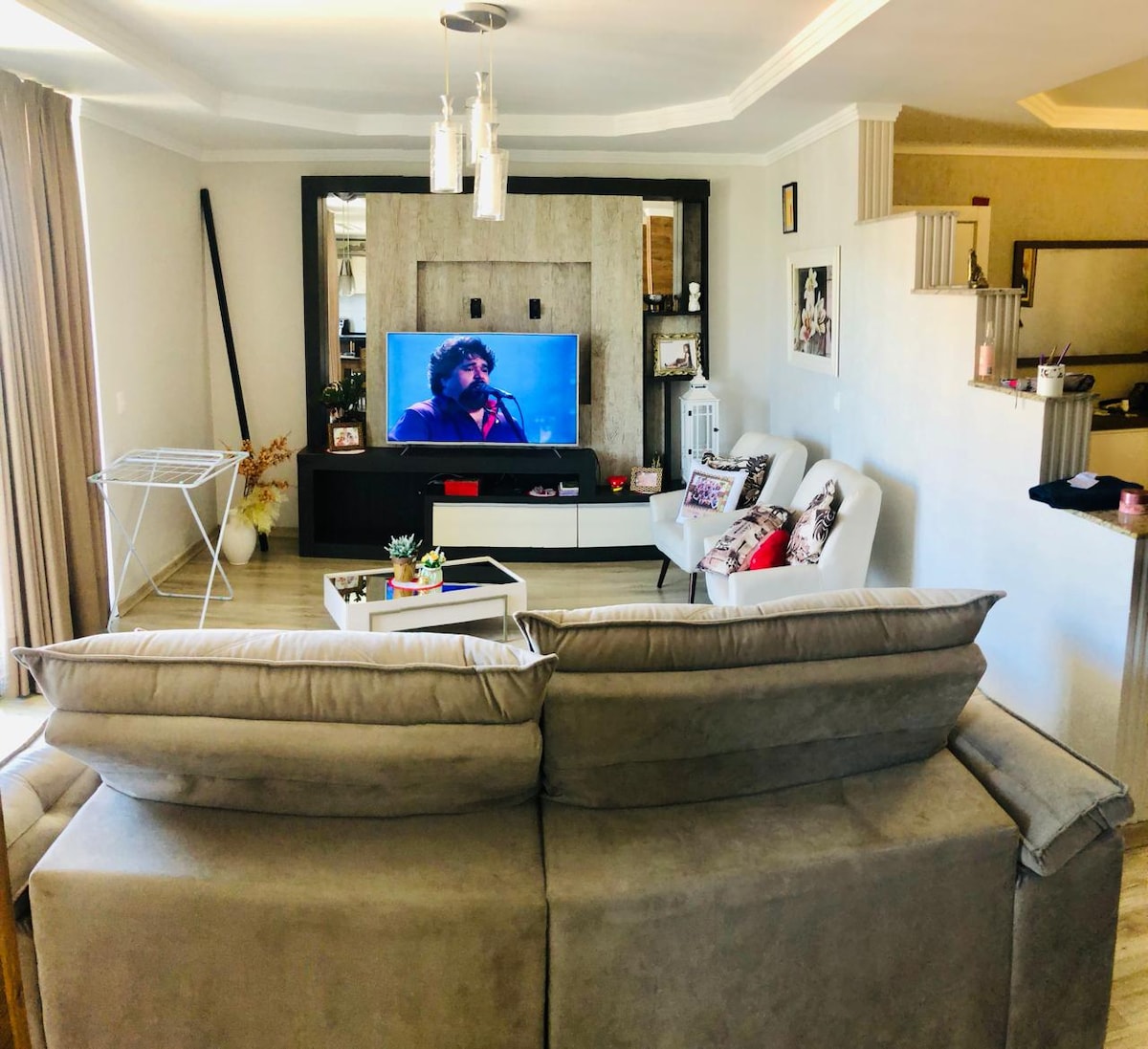 A comfortable living area is shown, featuring a large sofa with soft cushions facing a flat-screen television mounted on the wall. A light-colored coffee table rests in front of the sofa, accompanied by decorative plants. Natural light filters in through floor-length curtains, enhancing the inviting atmosphere.