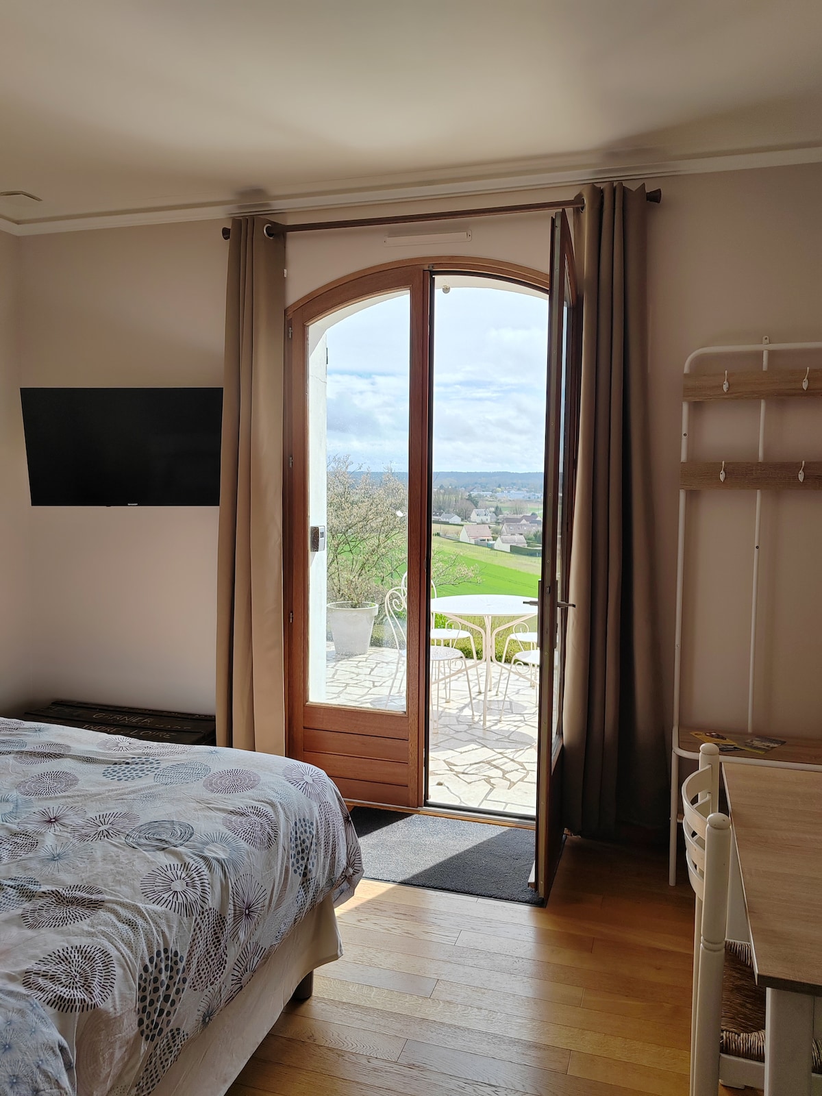 A spacious bedroom features a large bed with a patterned duvet cover. Natural light floods the room through an arched door opening onto a private terrace. A wall-mounted TV and wooden desk complete the space, while a scenic view of lush greenery is visible from the doorway.
