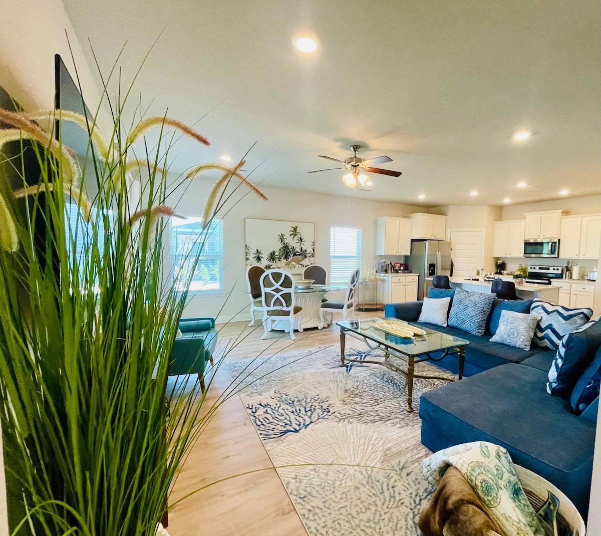 A spacious living area features a dark sectional sofa adorned with decorative pillows, ample natural light filtering through large windows. In the background, a dining set is visible, accompanied by a modern kitchen equipped with stainless steel appliances. A decorative rug adds texture to the floor.
