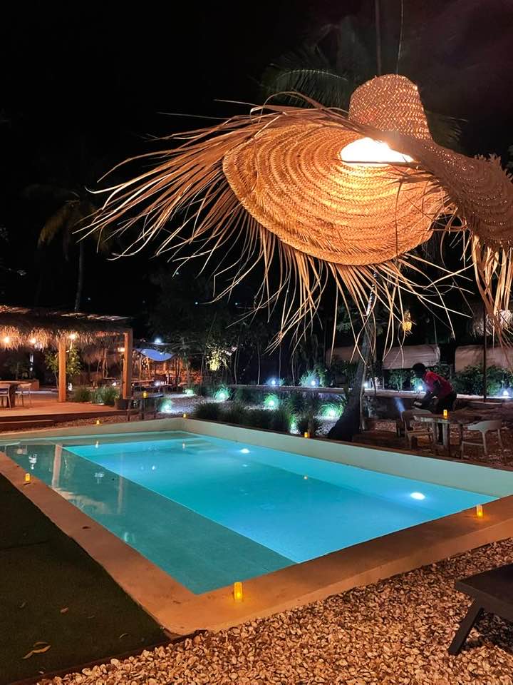 A tranquil pool is illuminated by soft lights, surrounded by lush greenery. A large straw light fixture hangs above, adding a decorative touch to the outdoor space. Nearby seating options are available, providing a relaxing atmosphere for guests to enjoy the serene environment.