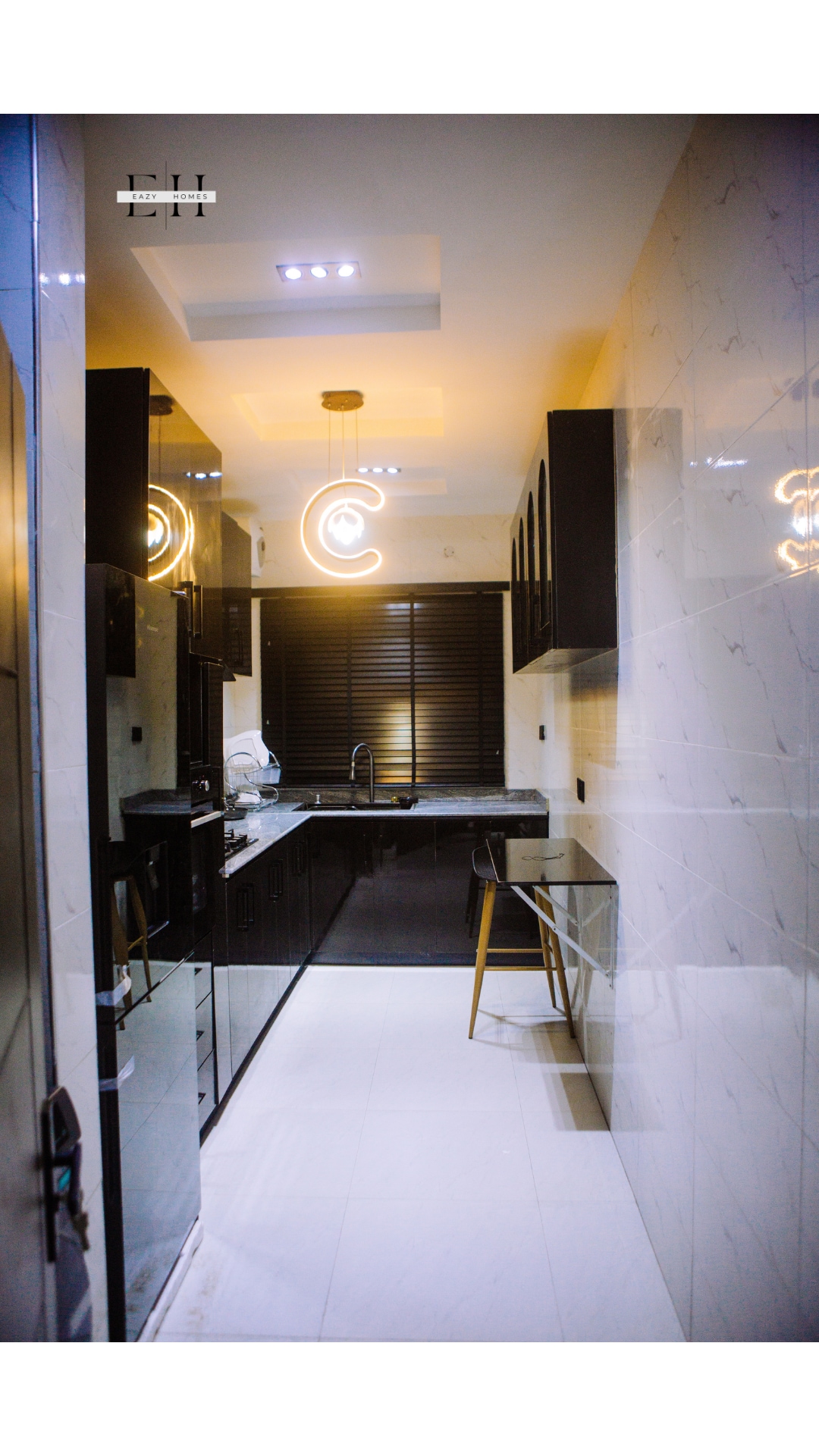 A modern kitchen is showcased with sleek black cabinetry and contrasting white countertops. Illuminated by stylish lighting fixtures, the space includes essential appliances and a small dining area presented by a tall stool at the counter. Natural light is filtered through the window with blinds.