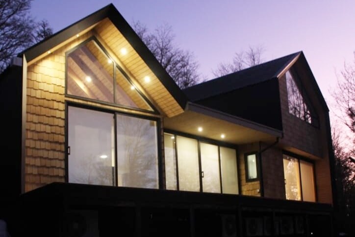 The exterior of a modern house is illuminated by soft lighting during twilight. Large glass panels provide views into the interior, while the sloped roof showcases a mix of wood and dark materials, creating a harmonious blend with the surrounding nature.