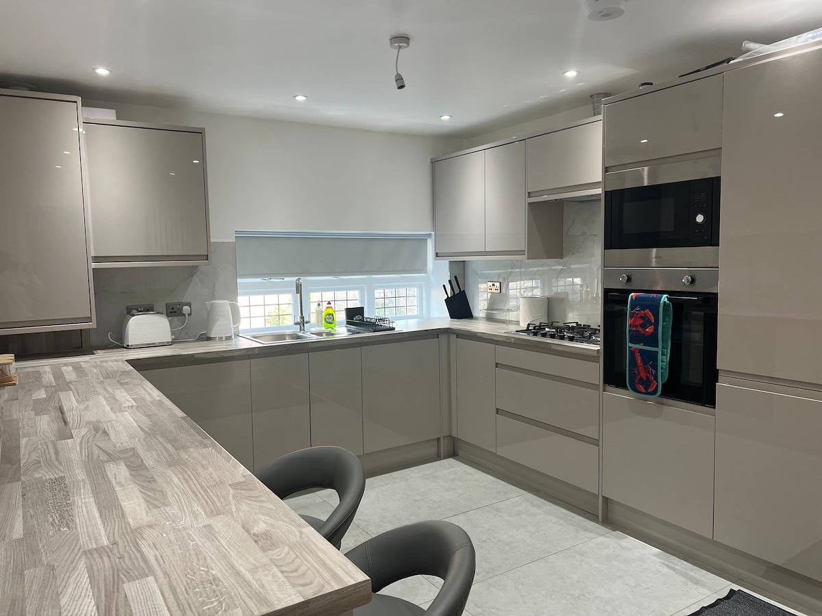 A modern kitchen features sleek, grey cabinetry complemented by a light-colored countertop. Stainless steel appliances, including an oven and stovetop, are prominently arranged. Natural light filters through a window above the sink, illuminating the spacious design. Two ergonomic chairs surround the kitchen island.