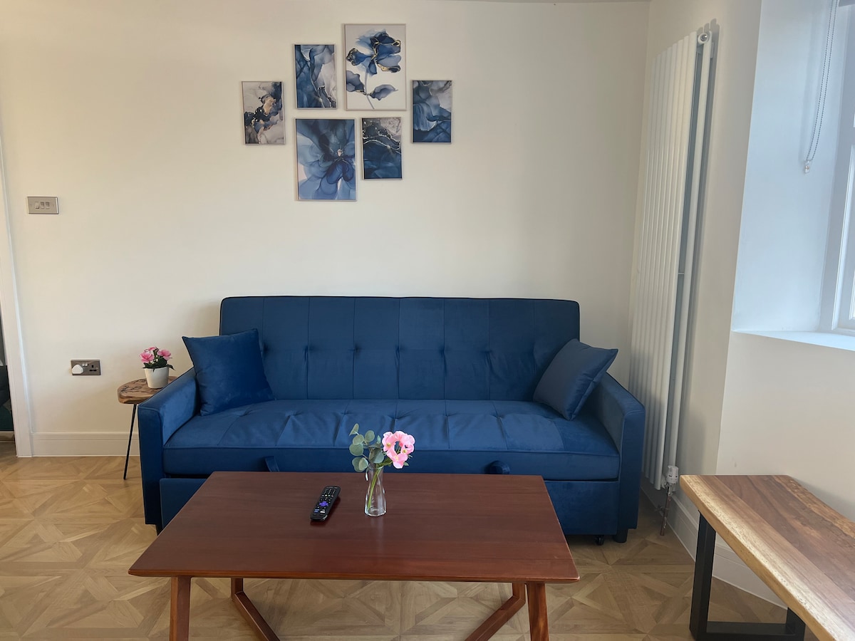 A comfortable blue sofa is positioned against a light-colored wall, complemented by decorative blue artwork above. A wooden coffee table with a remote control and a small vase containing flowers are situated in front of the sofa, enhancing the inviting living space.