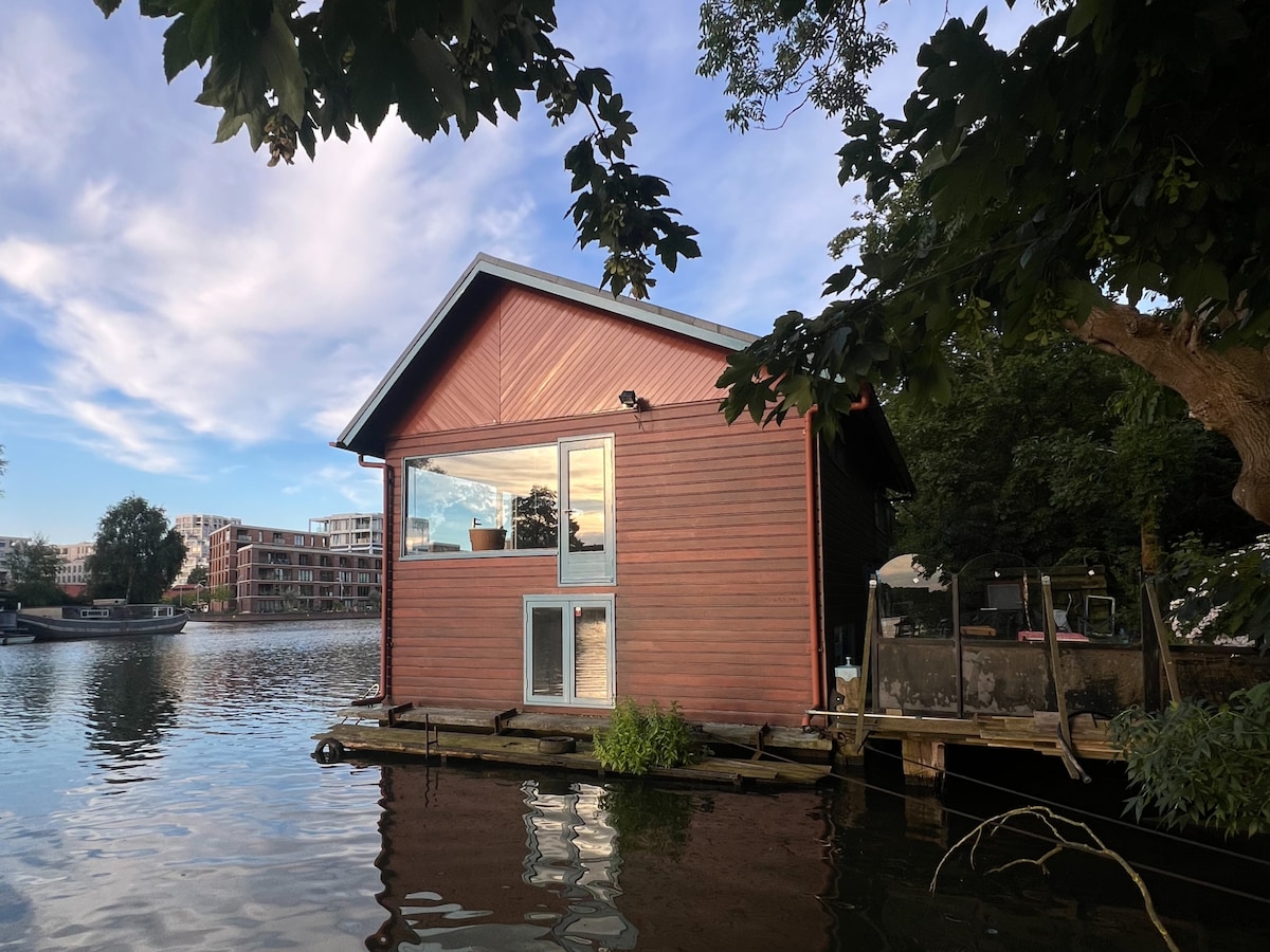 A wooden house stands on stilts above the water, surrounded by greenery. Large windows allow for natural light and provide views of the surrounding canal. The structure's vibrant red exterior contrasts with the blue sky and calm water, creating a tranquil setting.