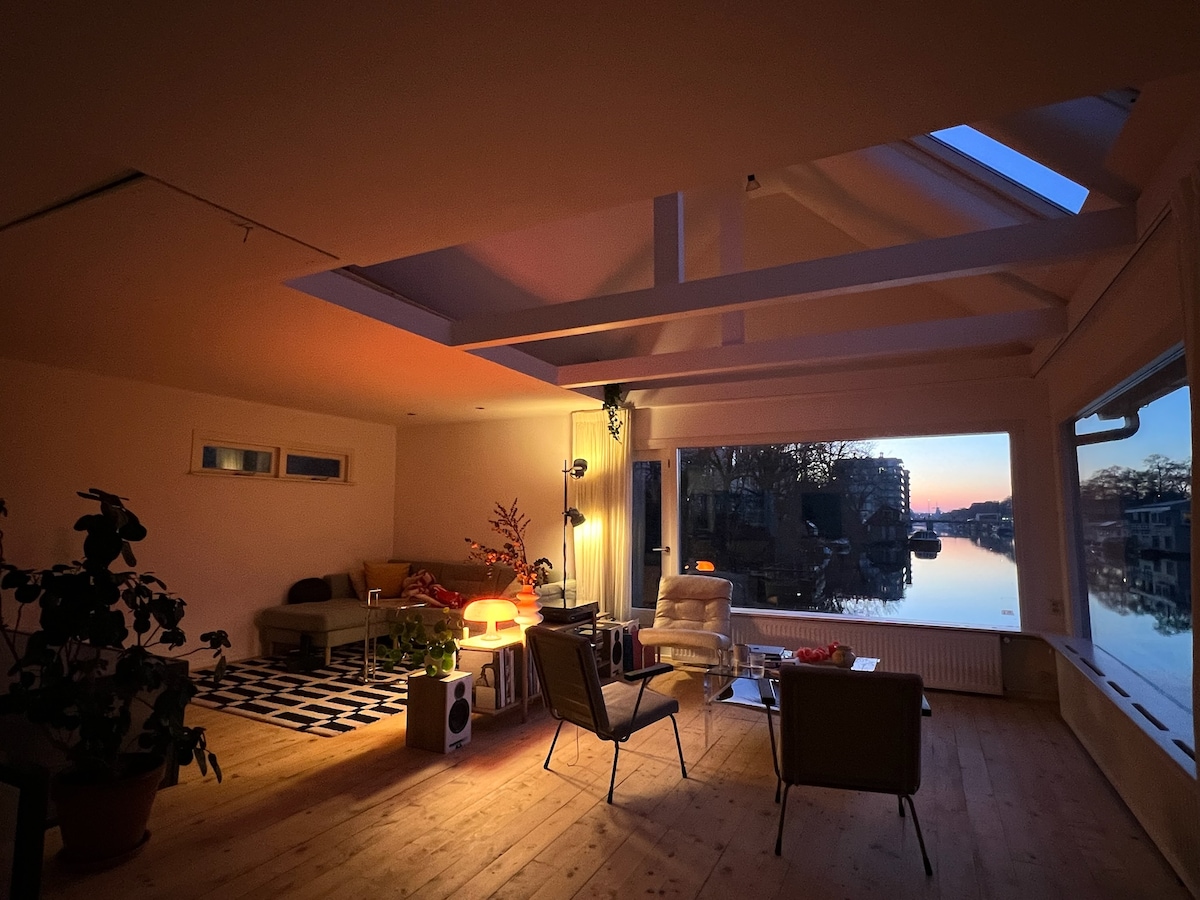 A spacious living area features large windows offering a view of the canal at sunset. Natural light filters through the skylights, illuminating the room. Comfortable seating arrangements include two chairs and a modern sofa, with potted plants enhancing the serene environment.