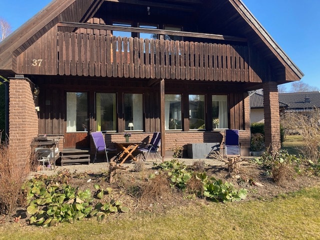 A two-story wooden house is depicted, featuring a broad balcony on the upper level. The front yard has a variety of plants and a gravel path leading to outdoor seating, including chairs and a small table. Natural light illuminates the large windows on the ground floor.