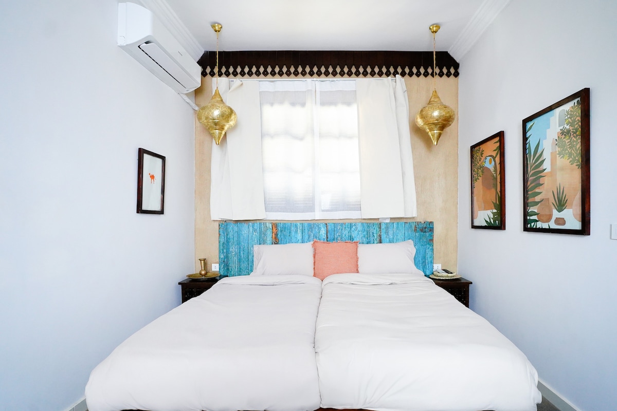 The room features two spacious beds with a light-colored linens set against a rustic blue headboard. Decorative gold pendant lights hang from the ceiling, complemented by artwork depicting nature on the walls. Natural light filters through sheer curtains, enhancing the tranquil ambiance.