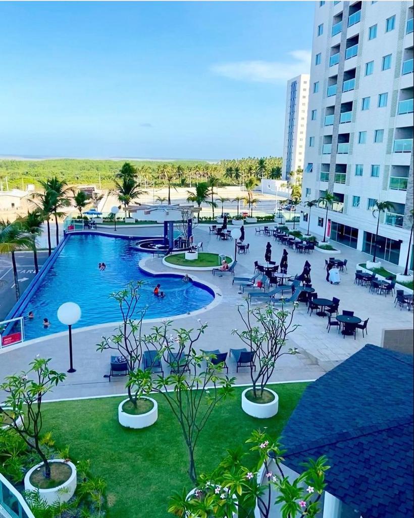 An outdoor pool is surrounded by lounging areas with tables and chairs, set amidst tropical greenery. Palm trees provide shade, and guests can be seen enjoying the sunny ambiance. The vibrant blue pool water reflects the clear sky, creating a refreshing atmosphere.