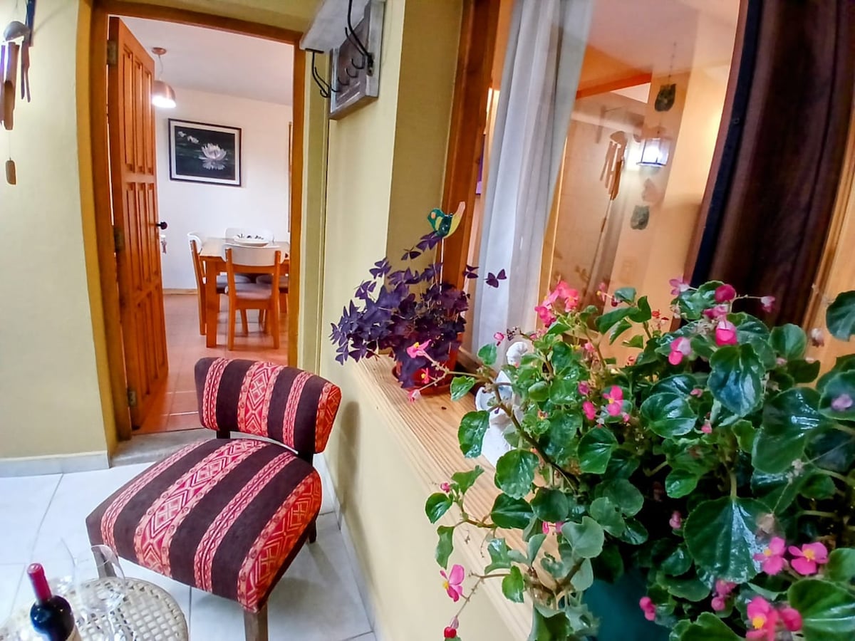 A comfortable seating area features a patterned chair beside a window adorned with vibrant plants. In the background, a cozy dining space is visible, furnished with a wooden table and chairs. Soft natural light enhances the welcoming environment.